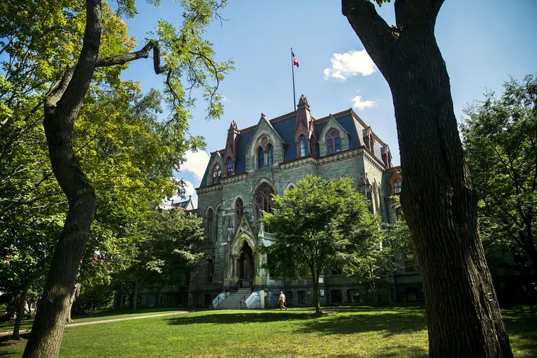 Wide shot of College Hall, a building with a facade of green, serpentine stone. A tall red and blue flag with the Penn crest is waving on top. 