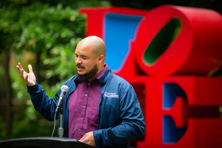 A male SPARC staff member in a blue sweater with a "Social Equity and Community" logo speaks in front of Penn's "love" statue.