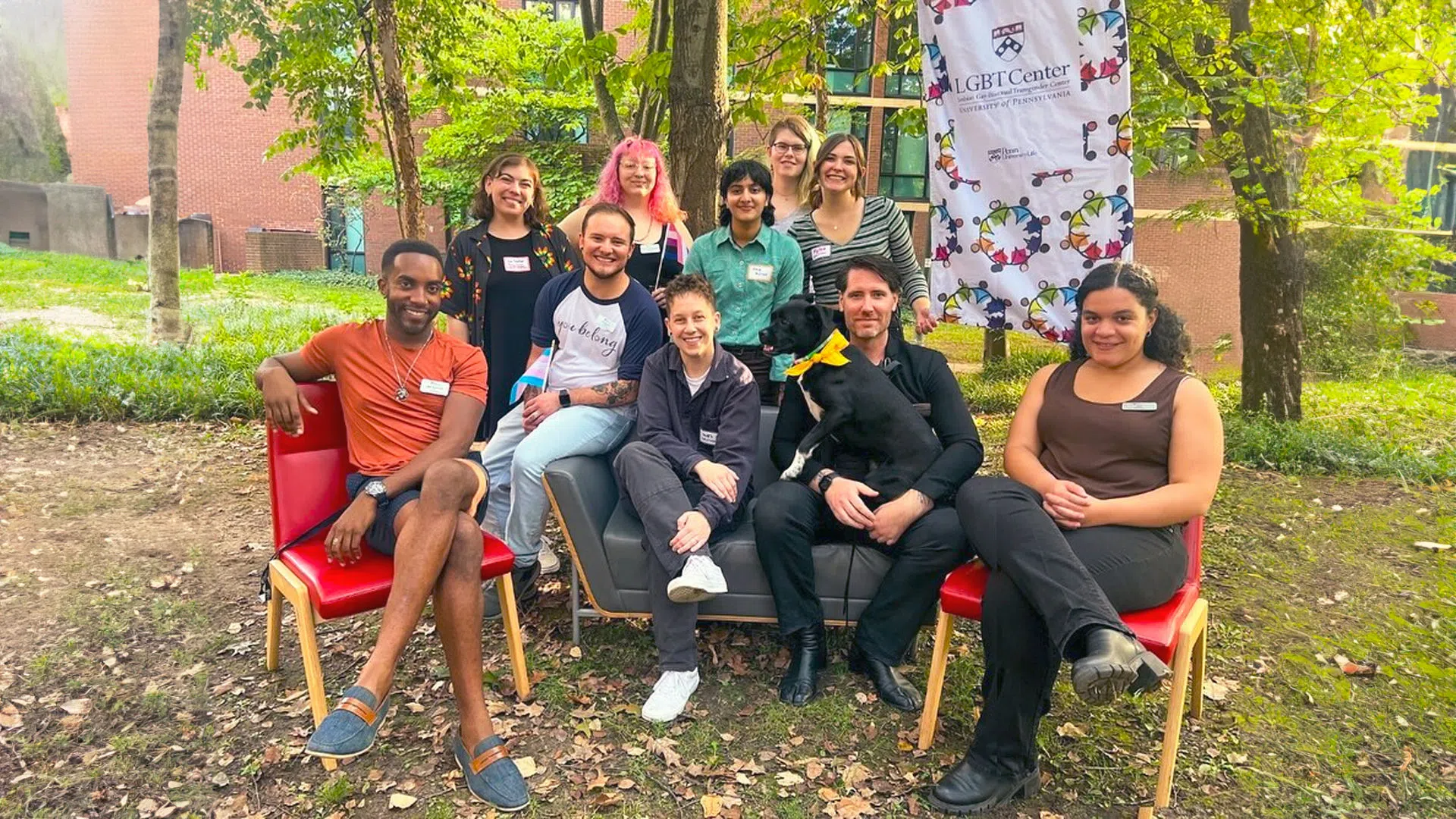 Center staff and student assistants pose for a photo, some seated, some standing. A staff member holds a dog, and they all wear pronoun nametags. One staff's shirt reads "You Belong."