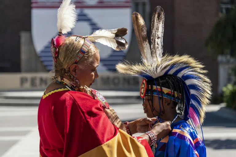 Elder & child, in tribal attire with colorful fabric, at Penn Commons for the Annual Pow-Wow. Elder helps child with clothing.