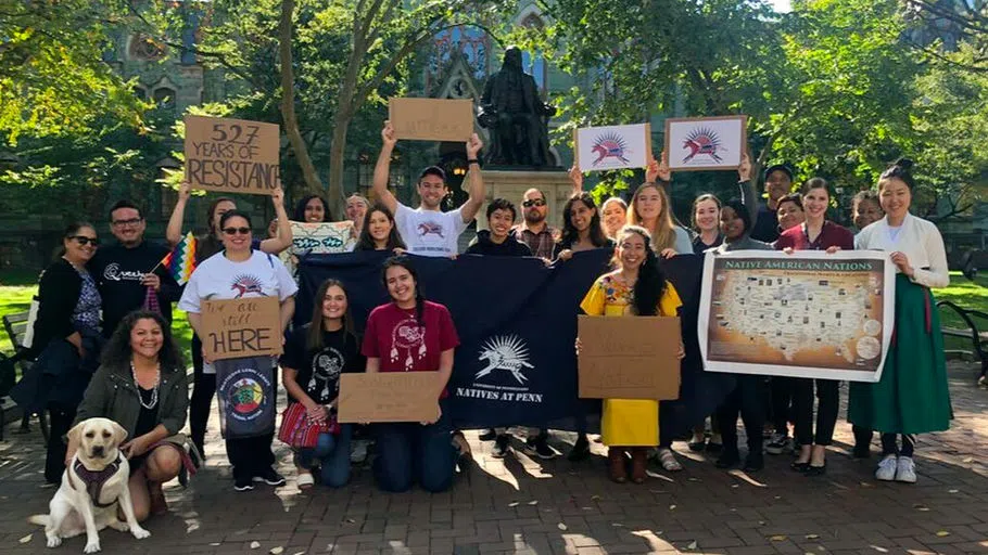 Staff, faculty, and students gather in front of the seated Ben Franklin statue at College Hall, holding flags, Natives at Penn logo, and signs saying "We are Still Here" and "527 Years of Resistance."