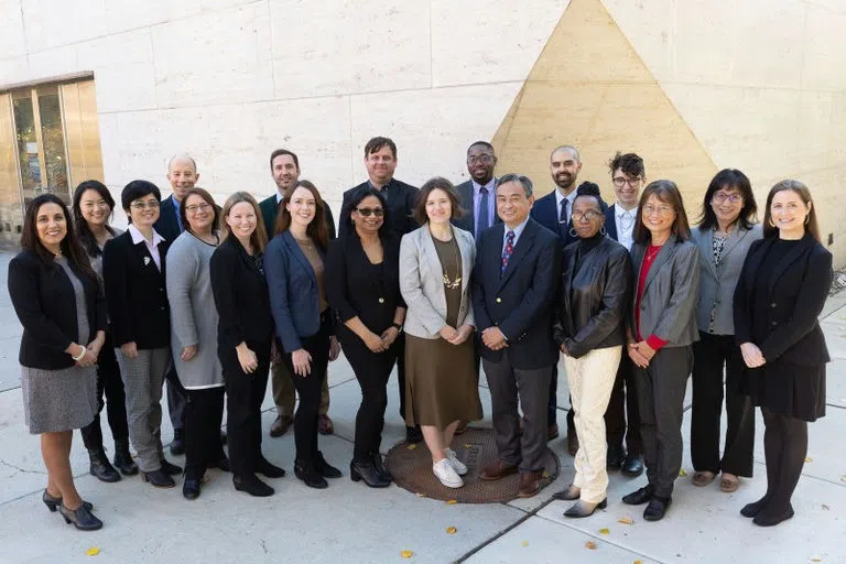 Nineteen students and staff in formal attire stand confidently smiling in front of the white façade of Perry World House, hands clasped together in front of them.