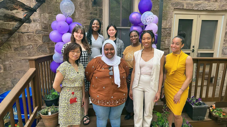 Three staff members and five students stand on the Center's back patio, all smiling, with purple balloon bundles behind them and potted plants at their feet.