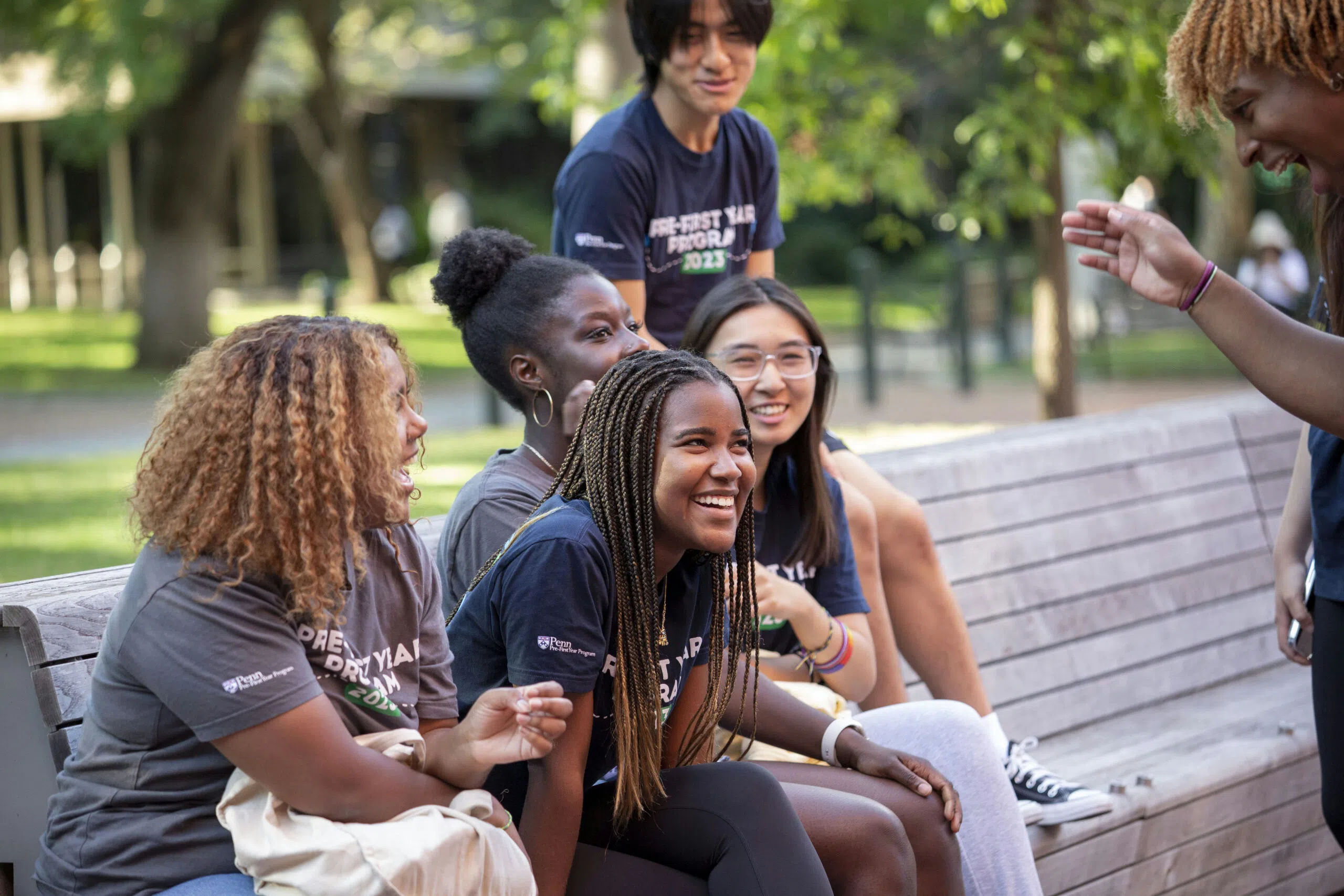 A group of six students are laughing and chatting on a bench. They are wearing navy blue and grey shirts that read "Pre-First Year Program 2023."