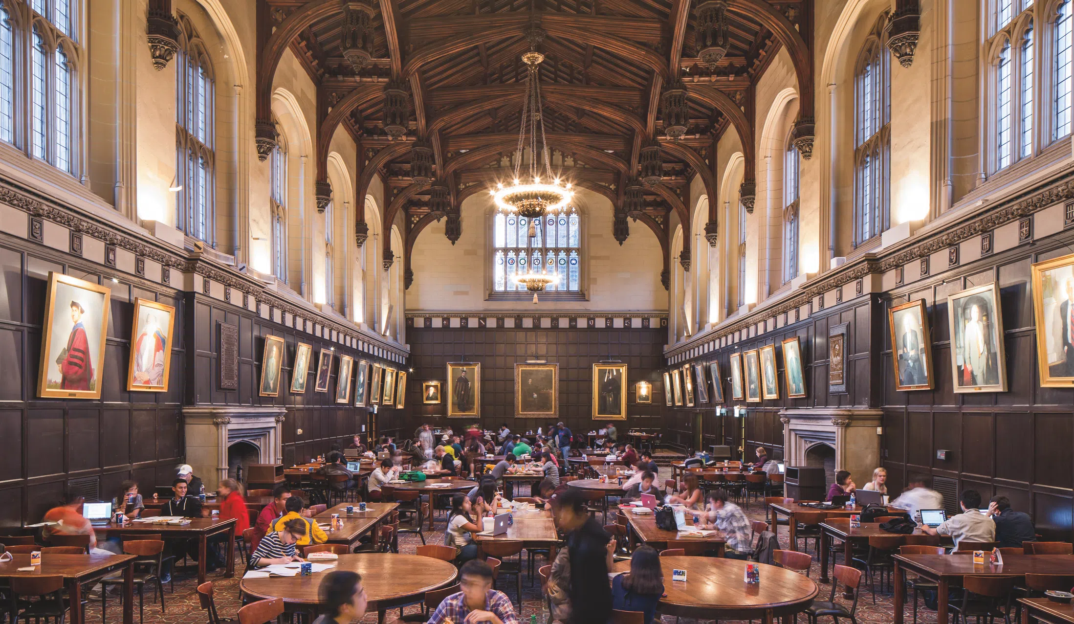 A large room filled with tables and students. Portraits line the walls.