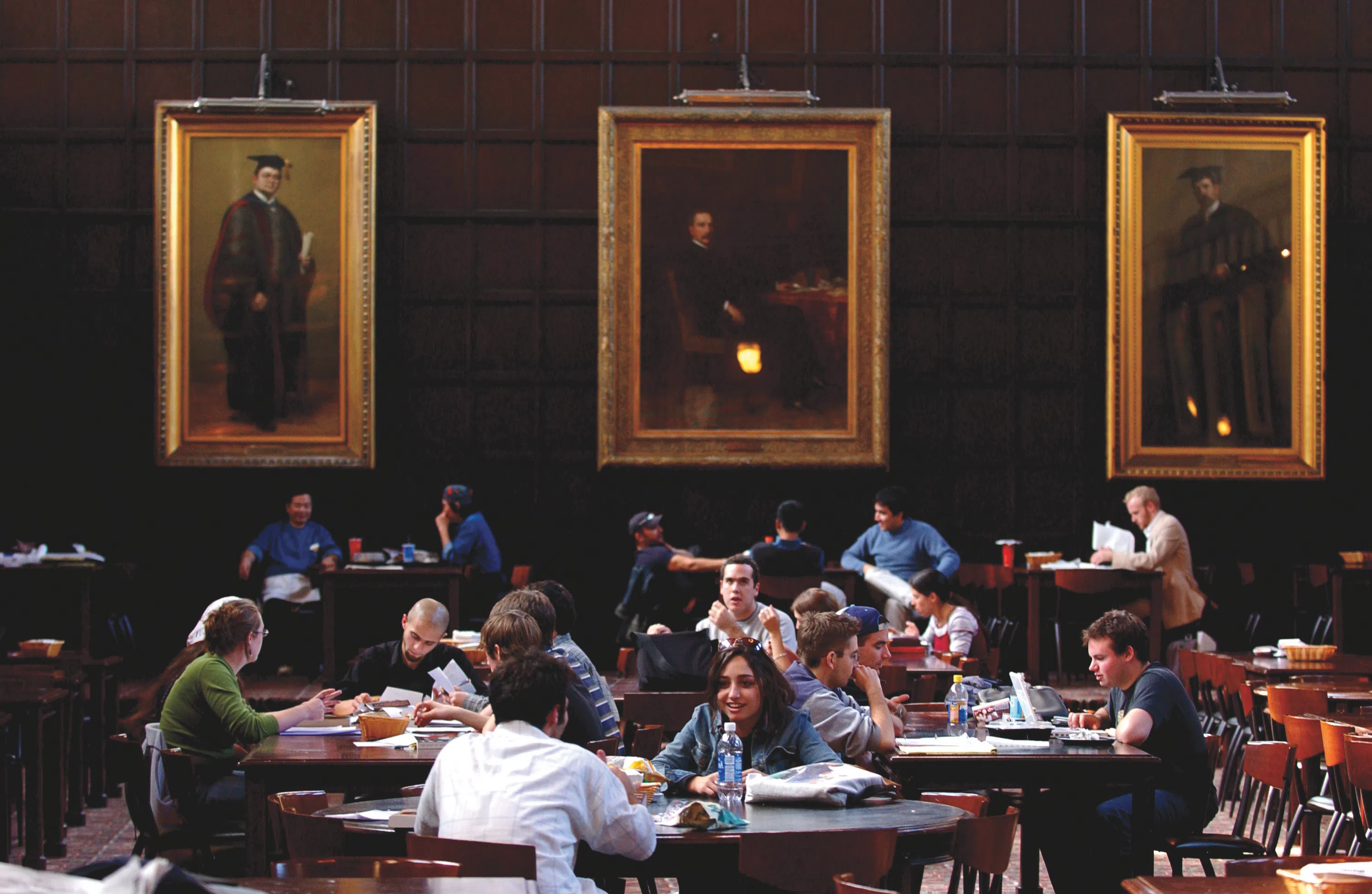 Students sit in a large room at tables eating and talking, large framed portraits hang in the background