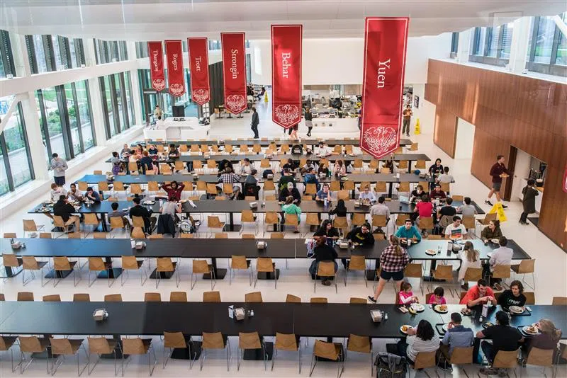 Students sit at long tables in a dining hall with red banners hanging above them 