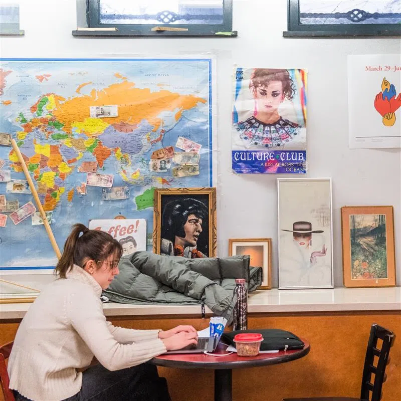 A student sits at a table in a cafe. Photos and maps are on the wall behind her