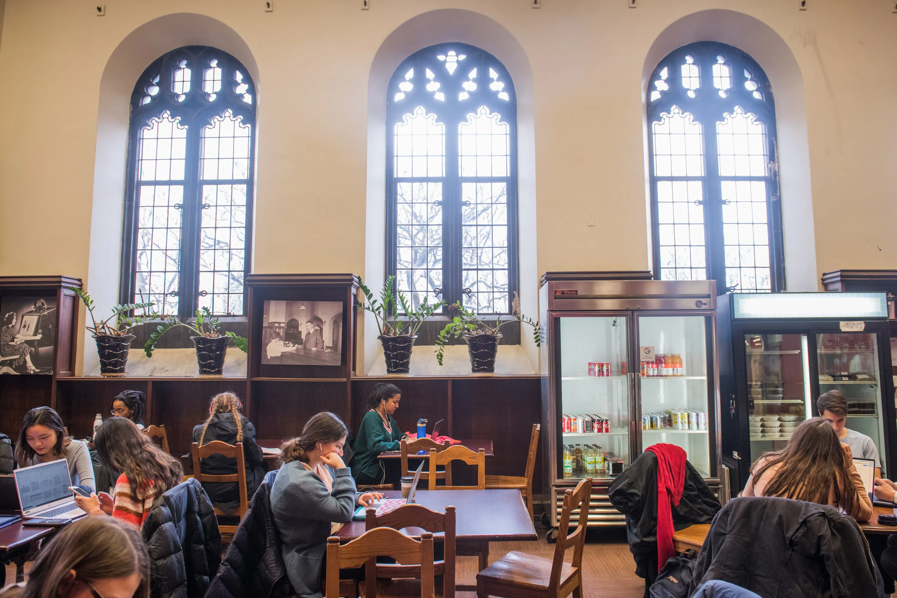 Students sit at tables at a cafe. Large gothic windows let in light. Vending machines are stocked with soft drinks.
