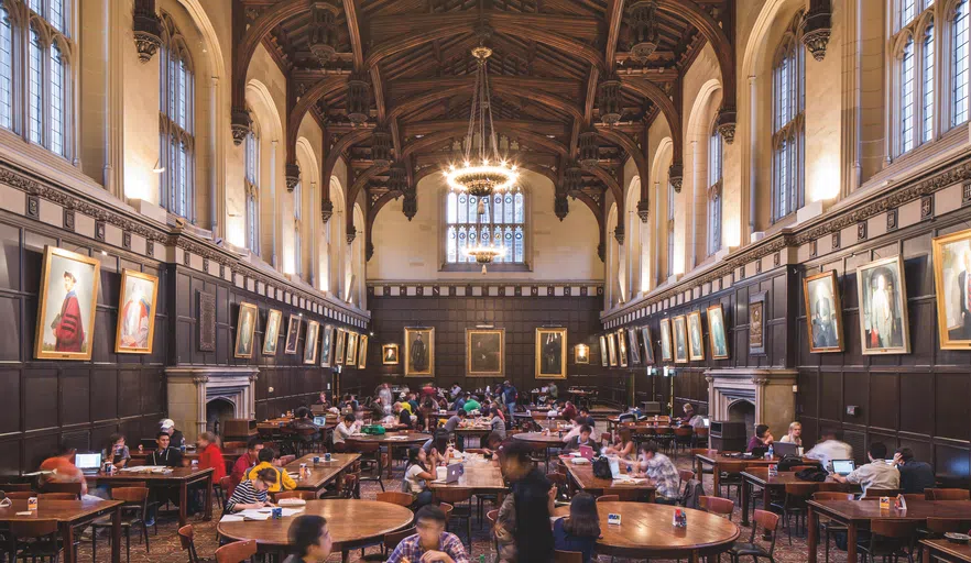 A large room filled with tables and students. Portraits line the walls.