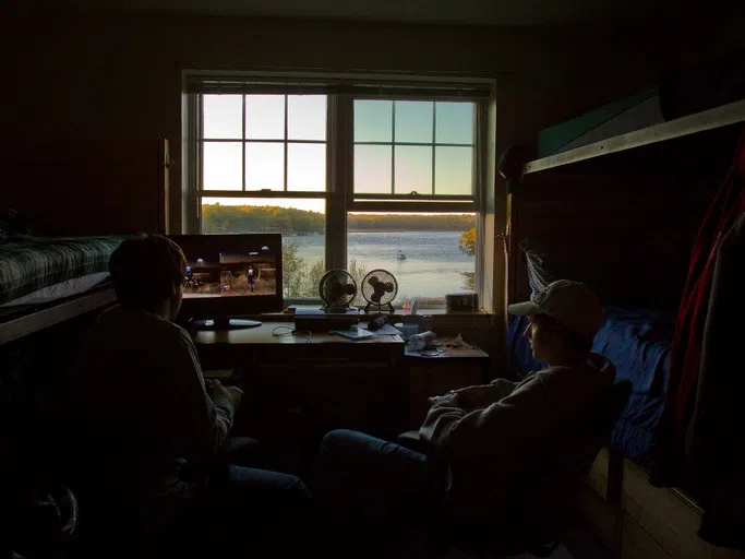 two students play a video game on a computer in front of a window that overlooks a sailboat on the water surrounded by trees