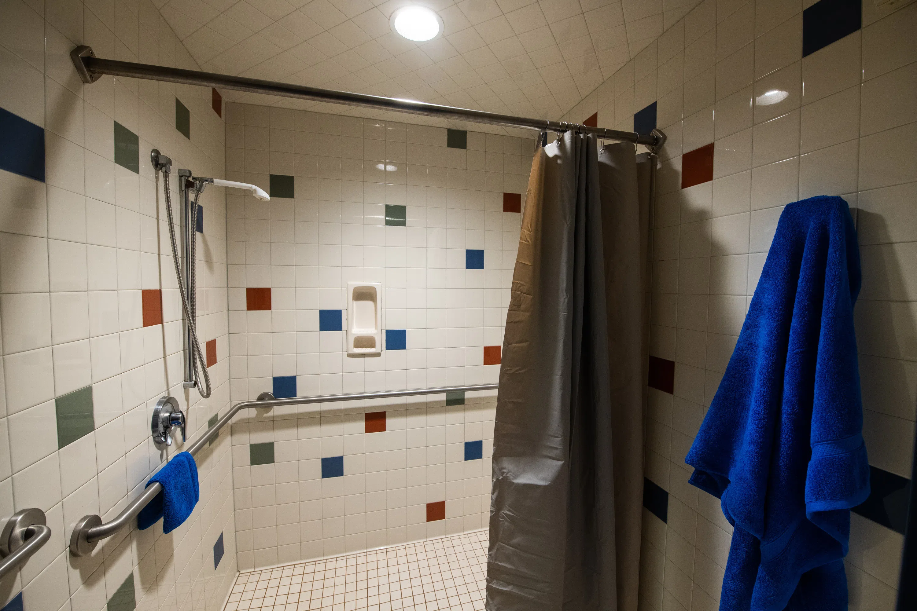 a large shower with railing and red, blue, green, and white tiles. A blue towel hangs from a hook adjacent to the shower.