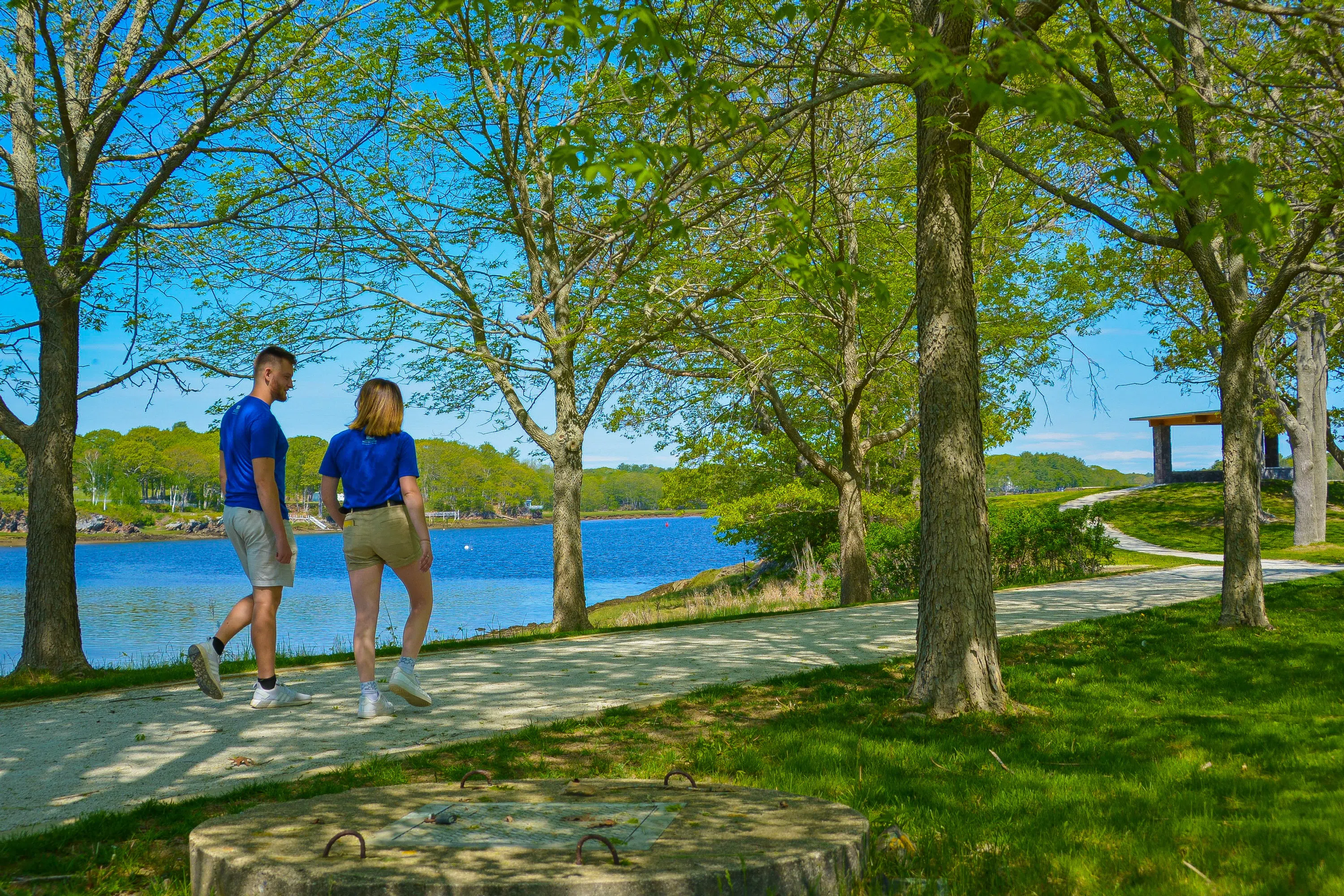two students walk up the winding path to the stone Kiosk, which is surrounded by trees and water on a peninsula..