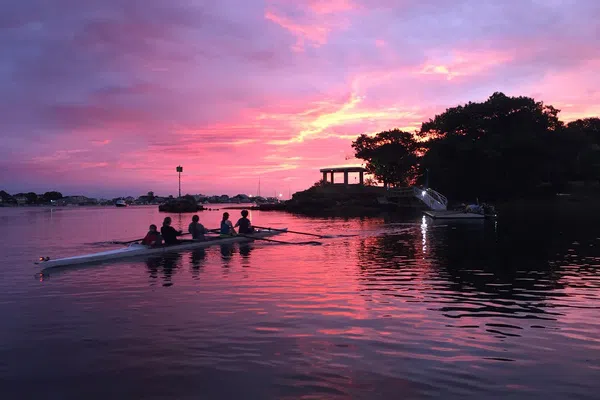 A crew team of five students in a racing boat row in the water towards a stone structure on a peninsula during sunset