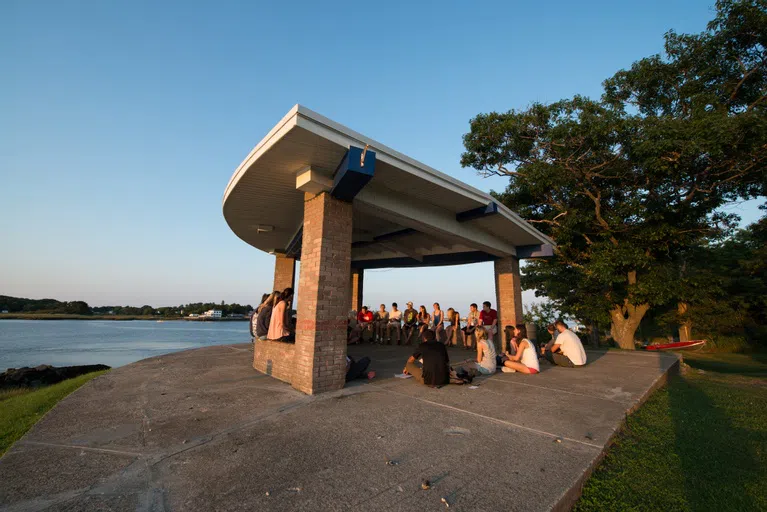 dozens of students sit o the ground of a stone kiosk surrounded by water and trees