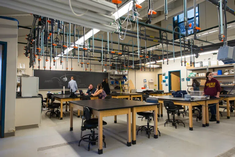 Large classroom with students sitting at lab tables and chairs and a professor writes on a chalkboard