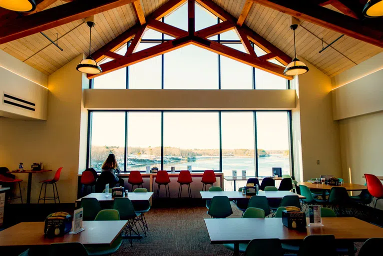 Large room with tables and chairs. A student sits eating in front of a wall of windows overlooking the water