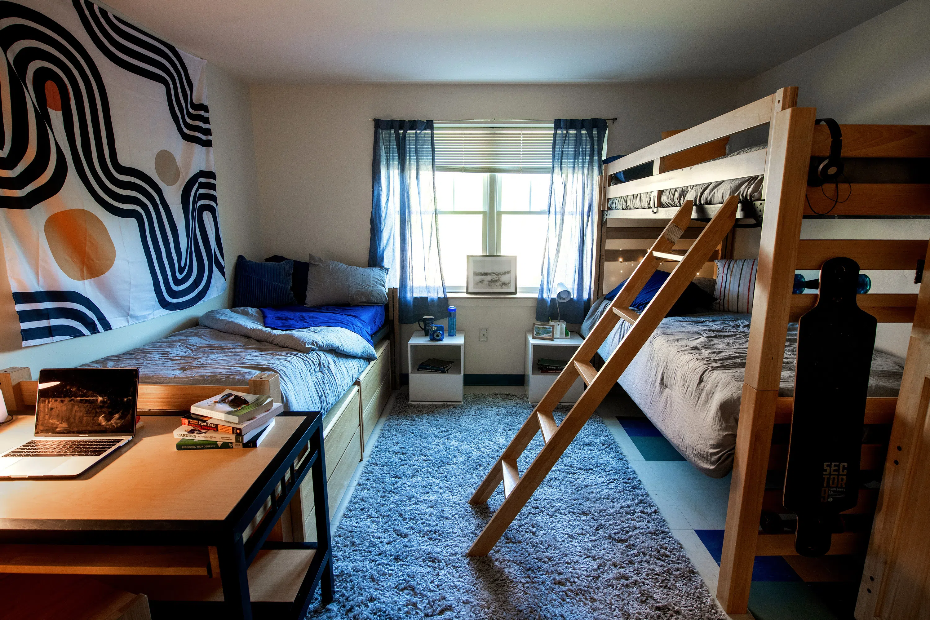 A single bed on the left of the room and the right side has a bunk bed with two beds. The back wall has a large double window. A desk is in the foreground. Decor includes: a tapestry, lamps, rug, books, and a long board.