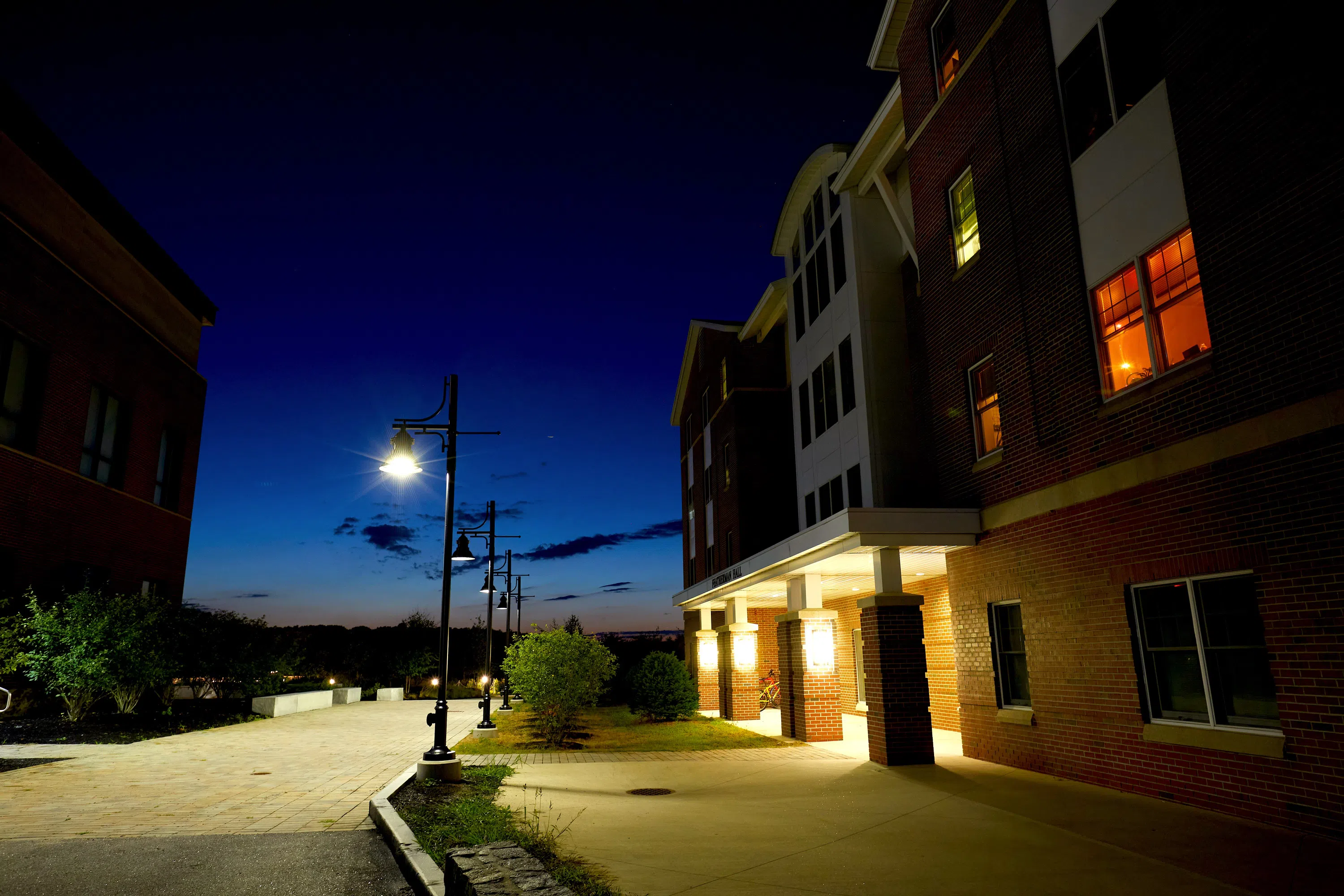 A brick building is illuminated with lights with stone walkway in front. Evening sky in the background