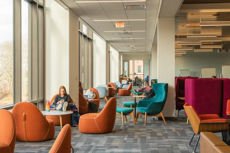 Students sit studying in bright large orange, teal, and grey chairs. A row of windows are on the left.