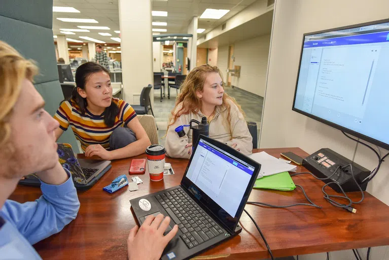 Three students looking at a laptop and computer screen on a wall.