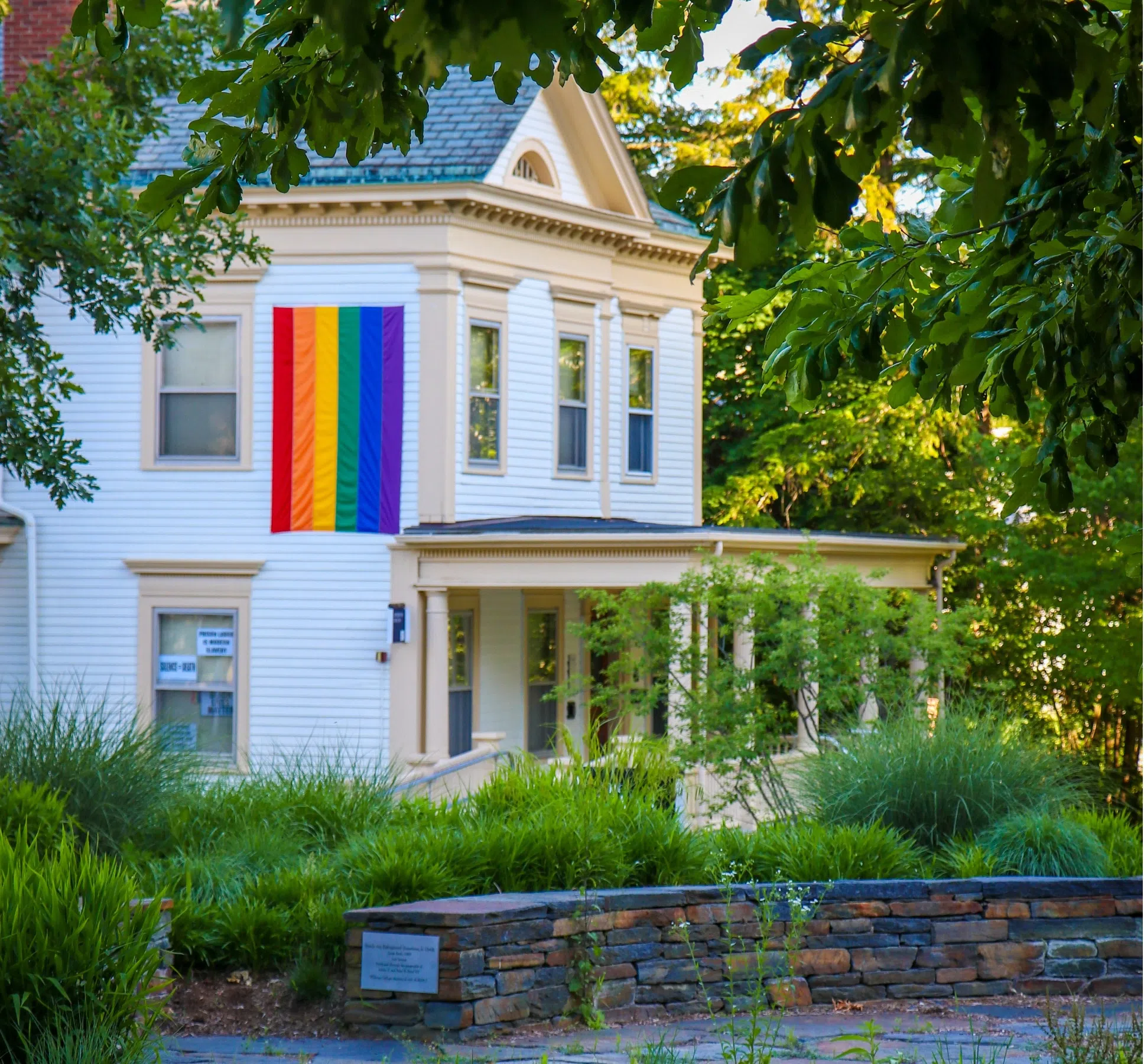 white house with pride flag hanging on the side, with trees and bushes surrounding it