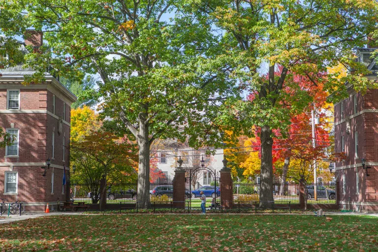 An image of a grassy quad leading up to the quad's entrance, where an arch and two red brick pillars connect Sage and Williams Halls. Fall trees cover the background.