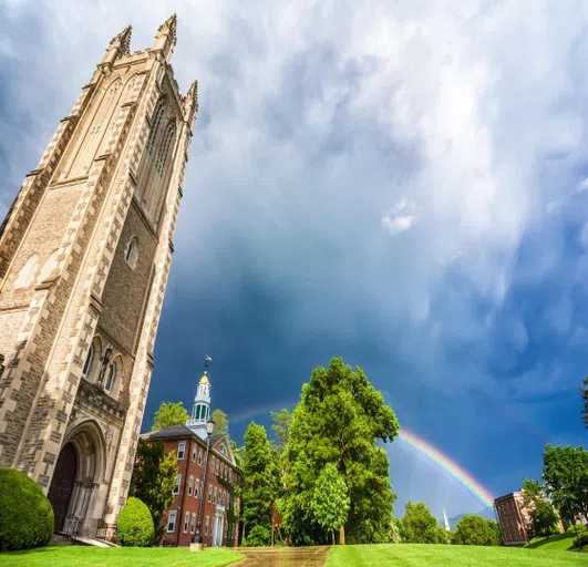 An image facing upward from a walkway at a rainbow, green trees and slightly cloudy skies, with the high peak of Thompson Chapel, a gray tower, looming above, and Griffin Hall, a red brick building with a small gold dome above, visible 